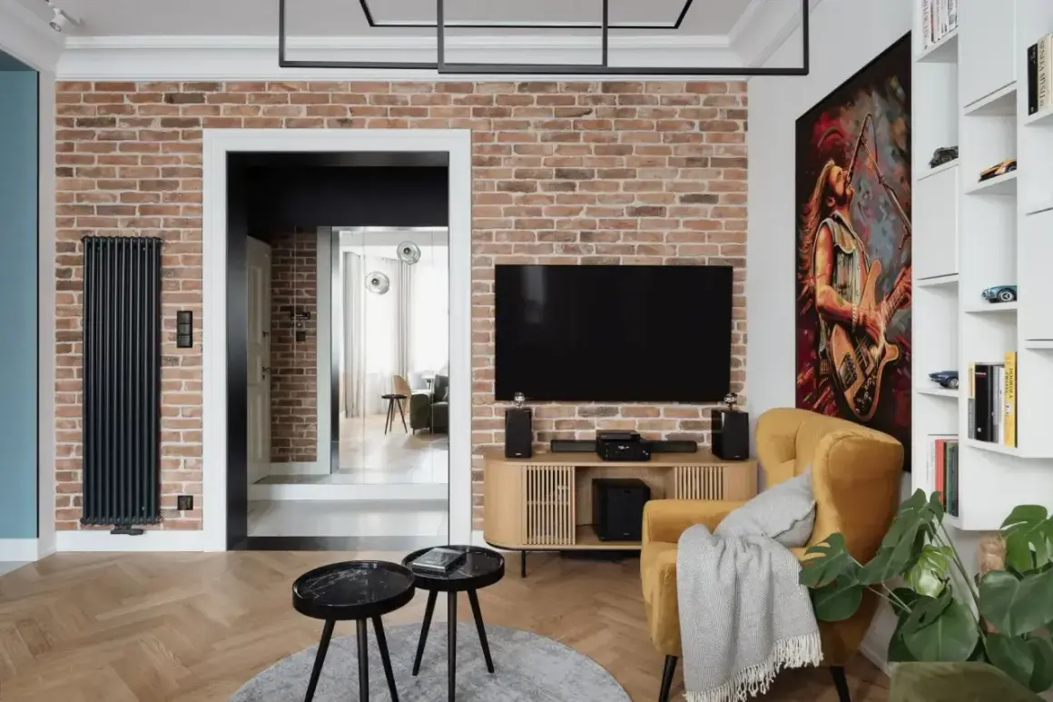 Nowoczesny salon z ceglaną ścianą, na której subtelne oświetlenie kamienia podkreśla jego fakturę. Obok telewizor i żółty fotel.