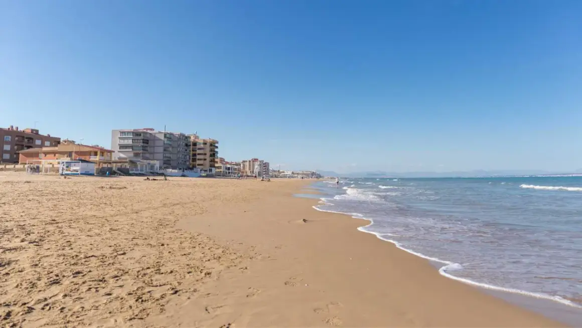 Arena dorada y edificios vacacionales en la playa Roqueta, con el mar Mediterráneo azul y olas suaves.