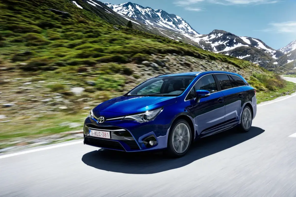 Niebieska Toyota Avensis T27 1.8 benzyna przemierza górską drogę, otoczona zielonymi zboczami i ośnieżonymi szczytami.