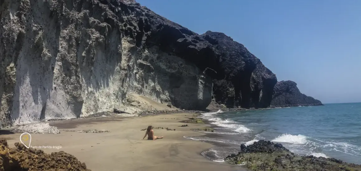 Una mujer descansa en la arena de la cala carbon, con imponentes acantilados de roca volcánica a su espalda.