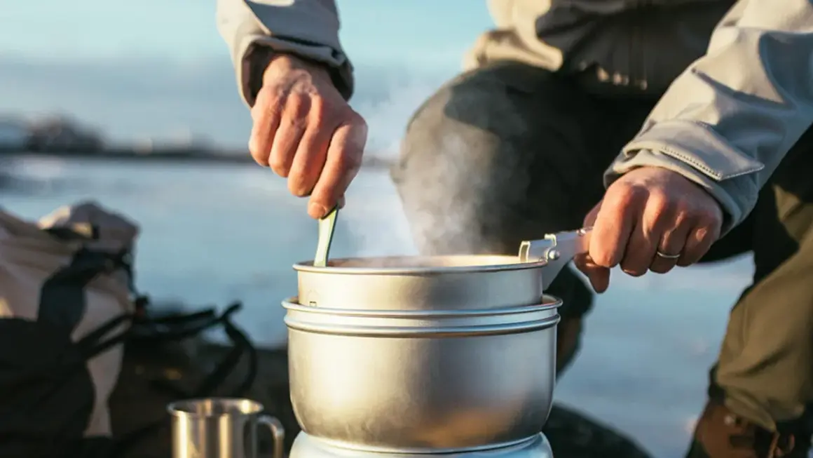 Person lagar mat utomhus, en del av mat vandring. Ångande kastruller och en mugg i metall.