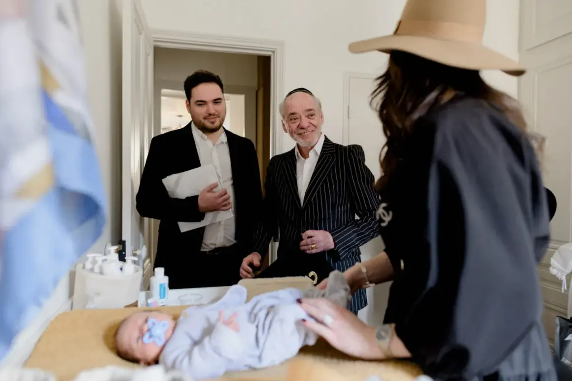 Un bébé est allongé sur une table, une tétine bleue dans la bouche. Des hommes en costume, dont un portant une kippa, observent le déroulement de la circoncision juive.