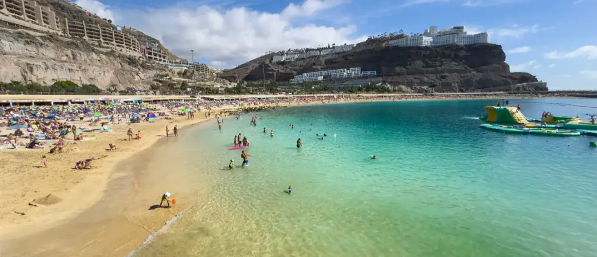 En livlig strand på Gran Canaria med solbadare, badande och en uppblåsbar vattenpark. En perfekt dag för att njuta av solen och havet.
