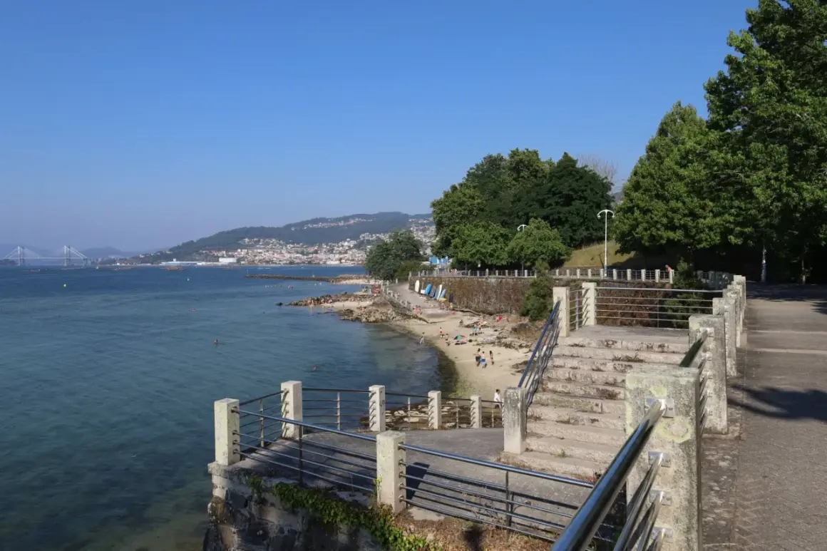 Vista de la playa de las barcas con gente disfrutando del sol y el mar. Al fondo, un puente y una ciudad en la ladera de una montaña.