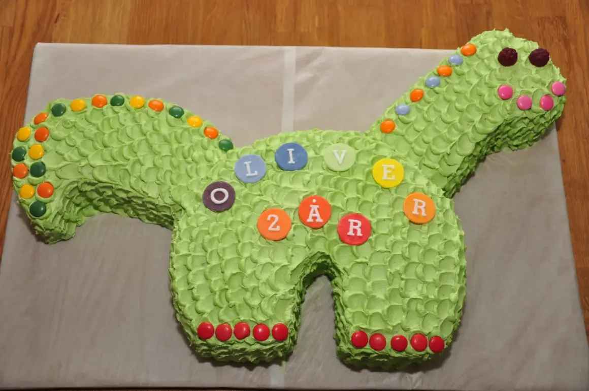 Enkel dinosaurietårta i ljusgrönt med godisdekorationer. Tårtan är formad som en dinosaurie och har bokstäver och siffror som bildar "OLIVER 2 ÅR".