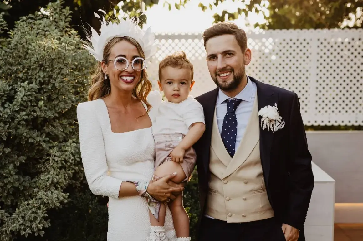Familia feliz en una boda. La novia, con un tocado blanco y gafas redondas, abraza a su hijo. El novio, con traje oscuro y chaleco beige, sonríe. Son los pajes boda más adorables.