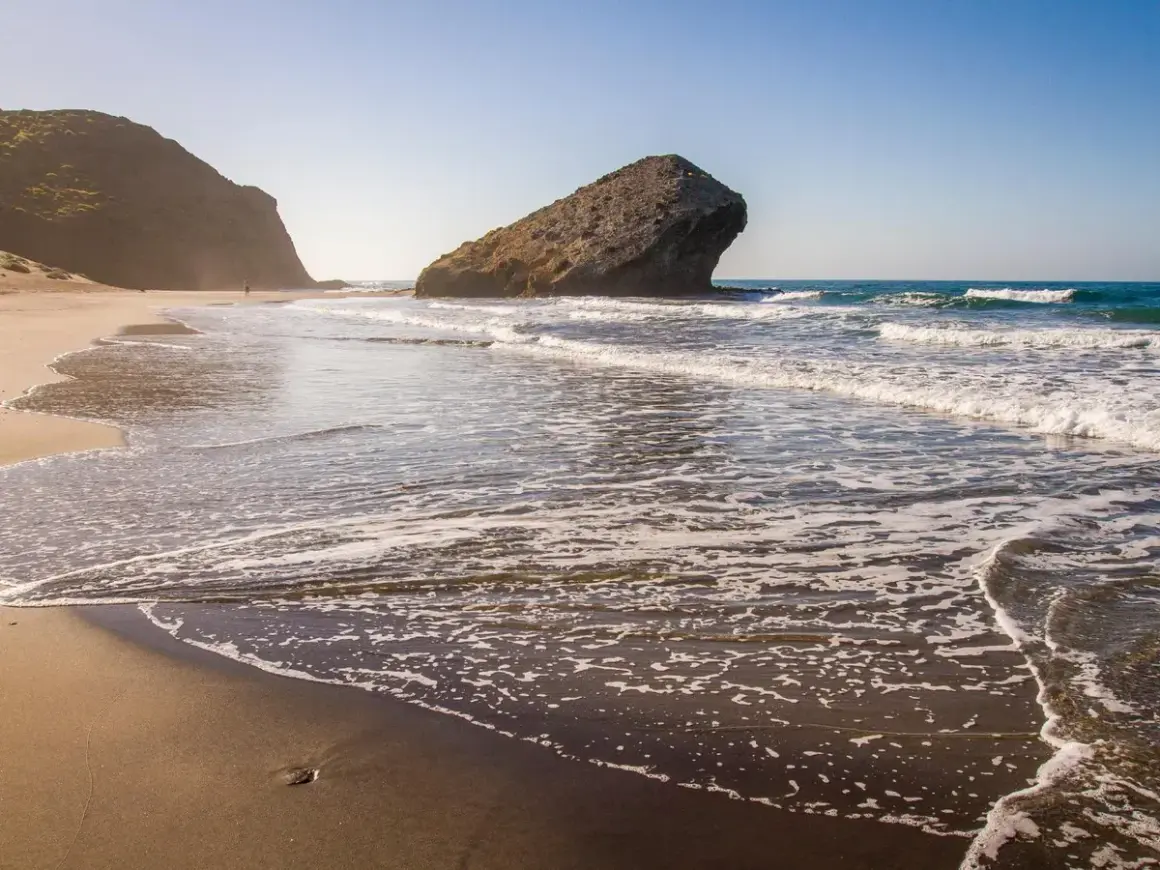 La duna de Monsul se extiende hacia el mar, con olas rompiendo suavemente en la arena dorada. Una figura solitaria camina por la playa.