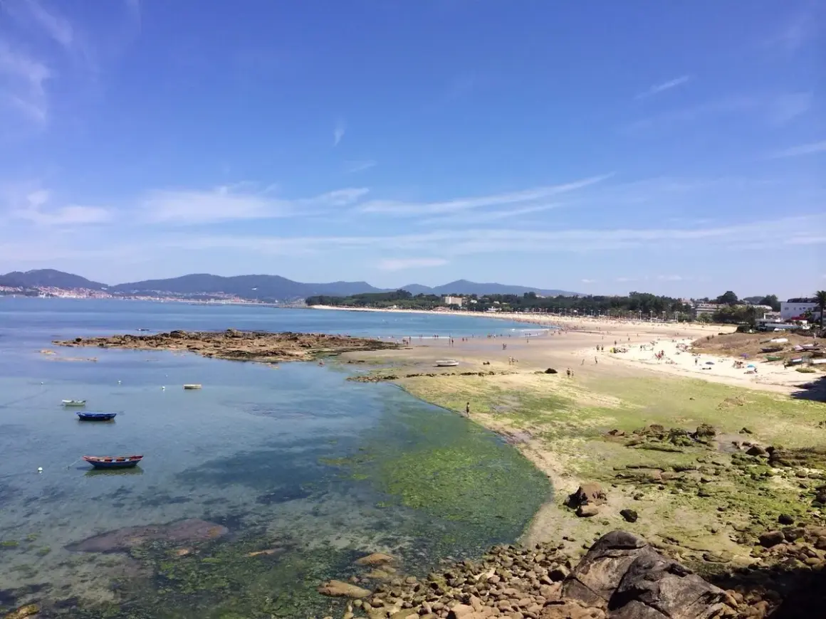 Vista de la playa de A Calzoa en Vigo, con gente disfrutando del sol y el mar. Barcas pequeñas flotan en aguas cristalinas.