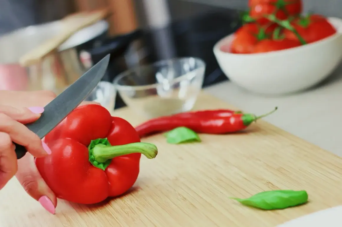 Ręka kroi paprykę czerwoną, bogatą w witaminy. Obok leży chili i pomidory, składniki pełne zdrowotnych właściwości.