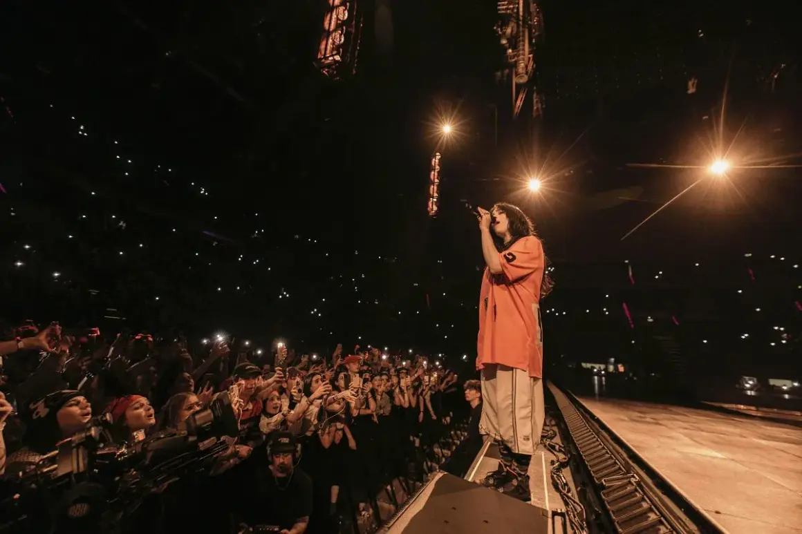 Billie Eilish śpiewa na koncercie przed rozentuzjazmowanym tłumem z telefonami w górze.
