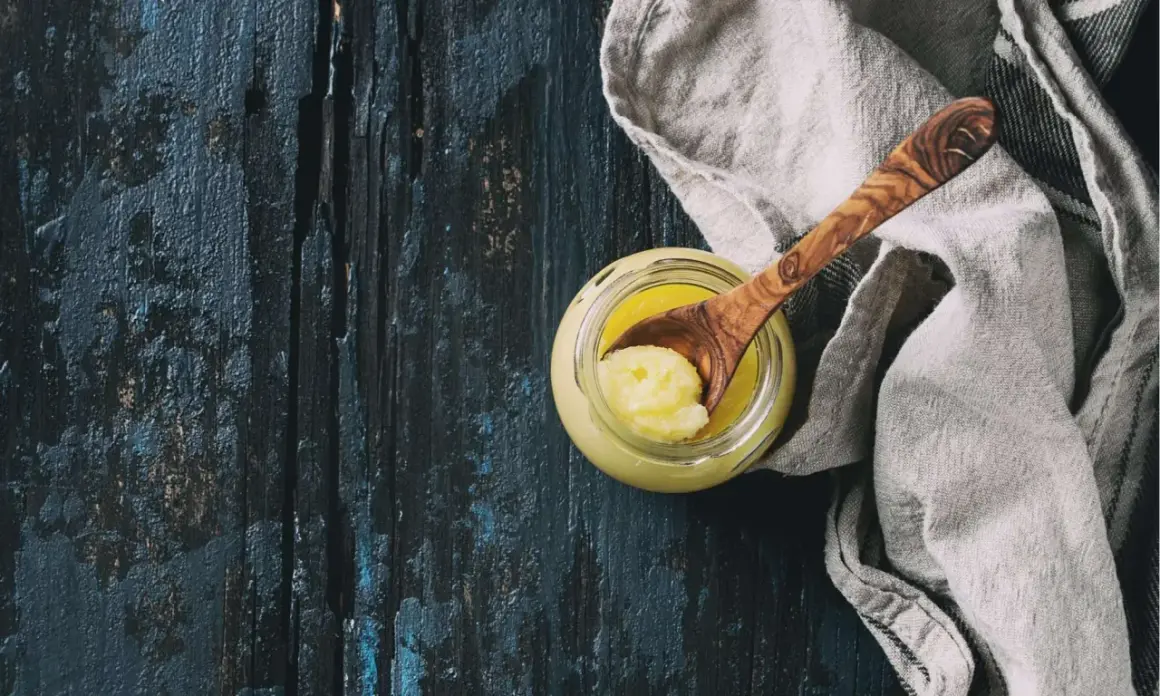 Ein Glas mit goldgelbem Ghee, daneben ein Holzlöffel mit einer Portion. Ghee ist geklärte Butter, ein vielseitiges Kochfett.