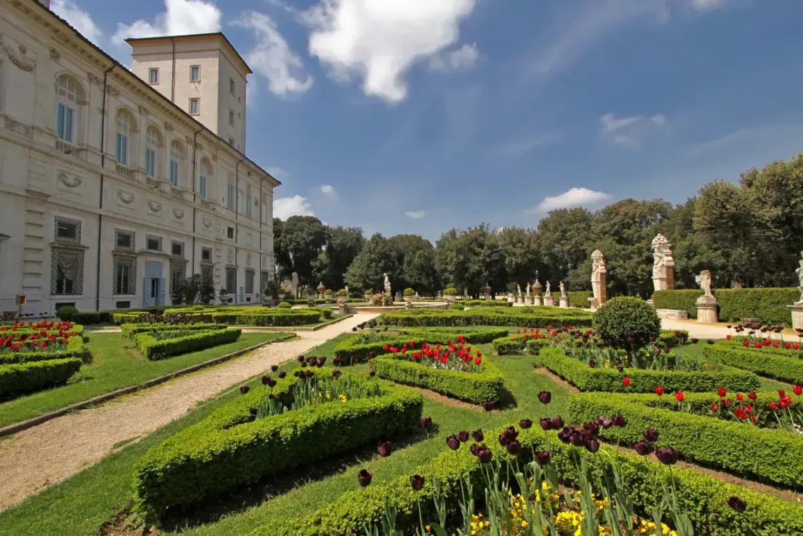 Giardini all'italiana di Villa Borghese con tulipani rossi e viola, statue e un palazzo sullo sfondo.