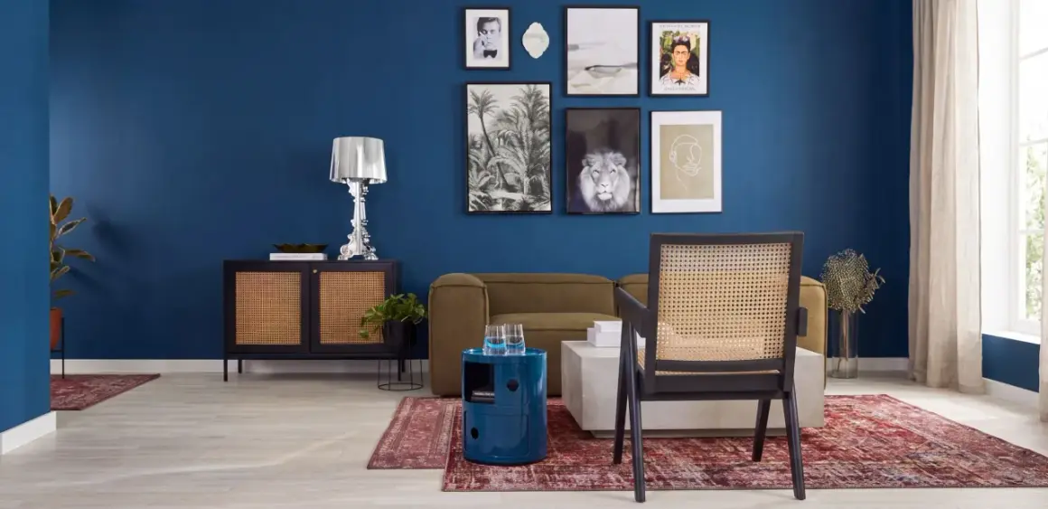 Ein stilvolles Wohnzimmer mit einer tiefblauen Wand, die mit Kunstwerken geschmückt ist. Ein Rattan-Stuhl steht vor einem Sofa, daneben ein runder, blauer Beistelltisch.