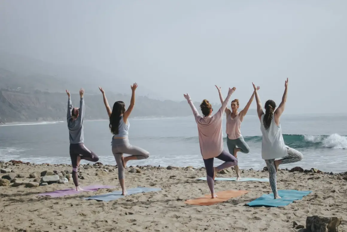 Cinco mujeres practican poses de yoga para 4 en la playa, con los brazos levantados hacia el cielo.
