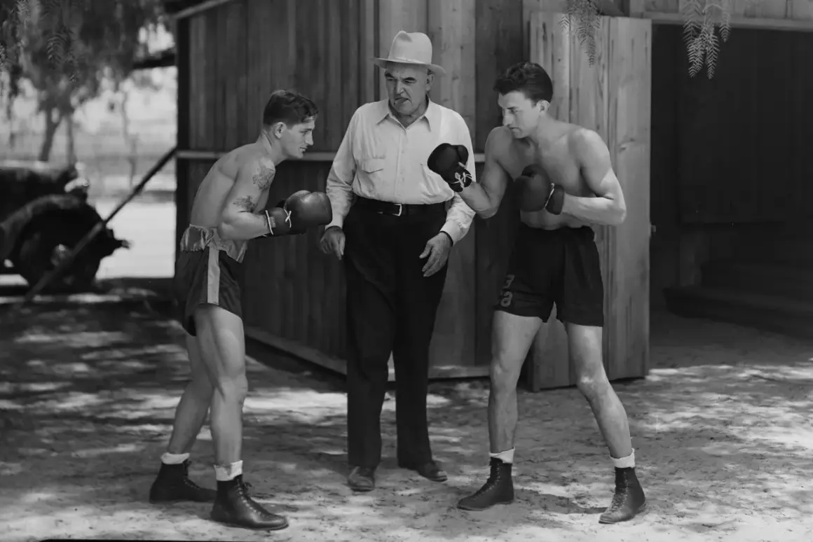 Two boxers spar while a trainer watches. The boxer on the left has a tattoo. This scene evokes the spirit of legendary boxer James J. Jeffries.