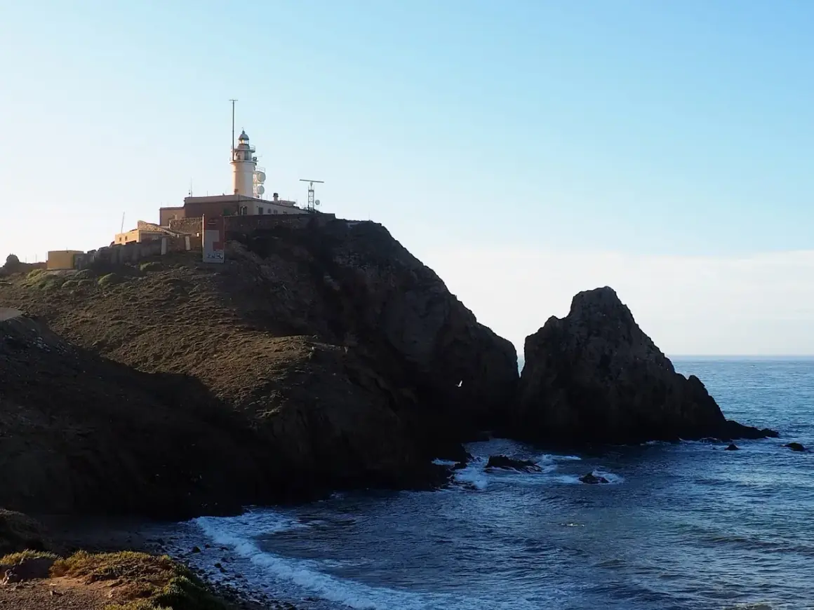 Faro en la cima de un acantilado rocoso sobre la cala del corralete. El mar azul choca contra las rocas.