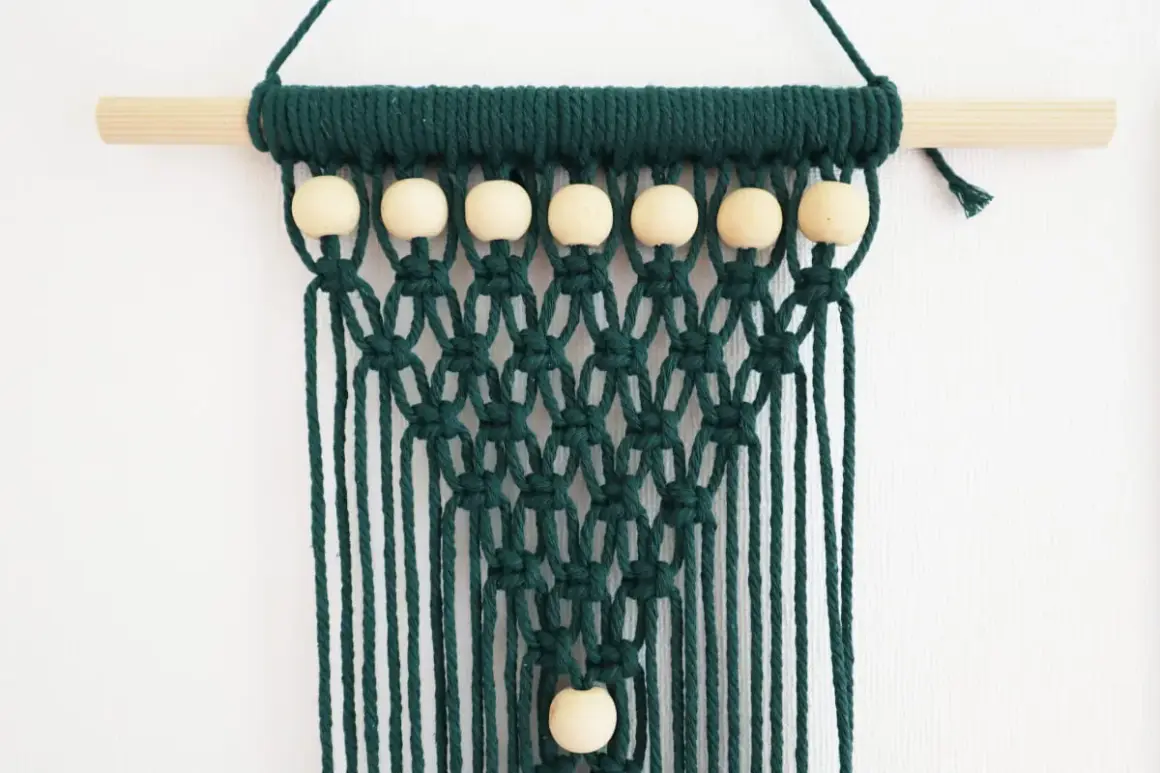 Tenture murale en macramé vert foncé avec perles en bois sur une tige en bois.