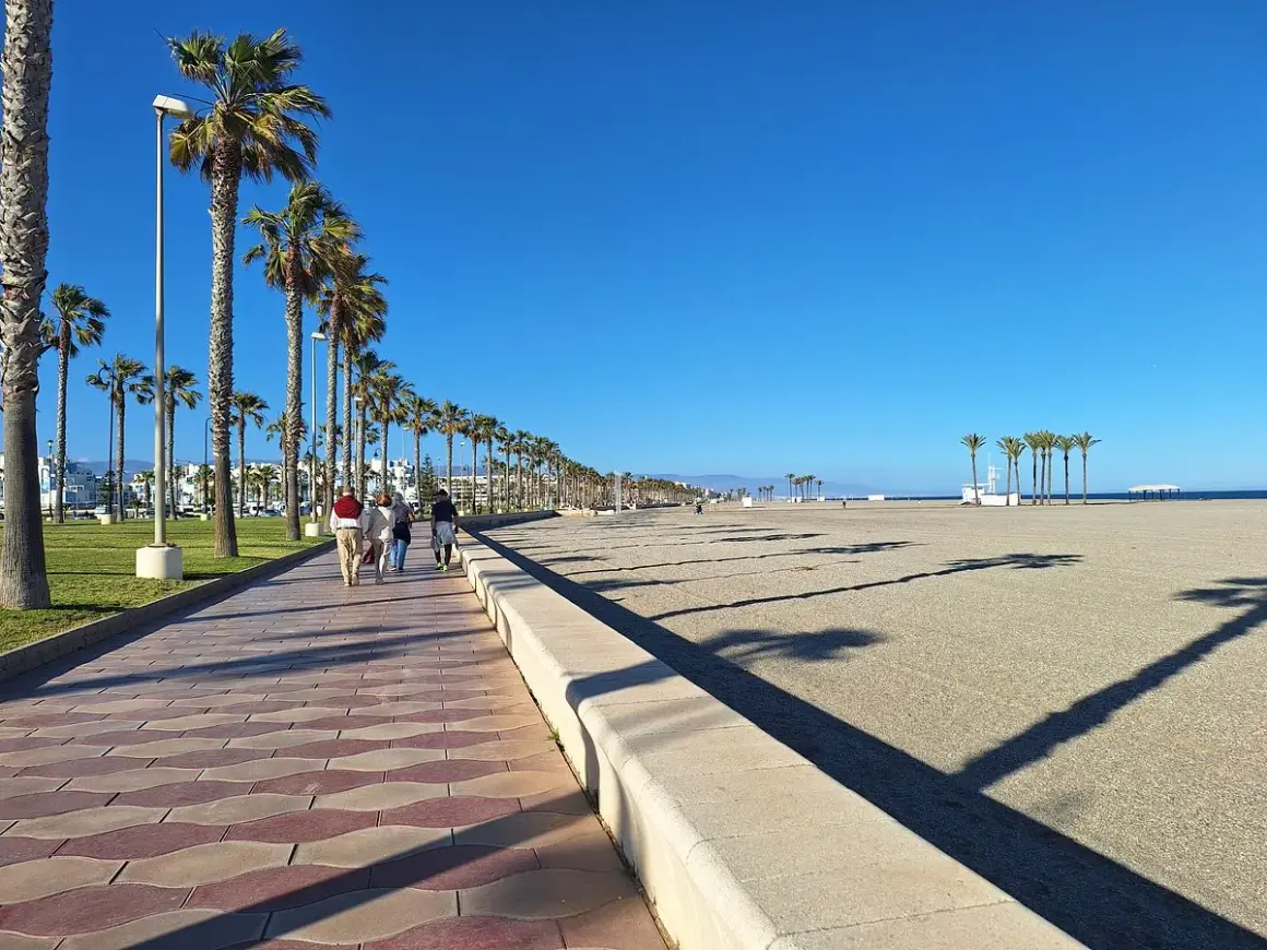 Paseo marítimo con palmeras en el **parque Roquetas de Mar**. Gente camina por la senda adoquinada junto a la playa de arena bajo un cielo azul.