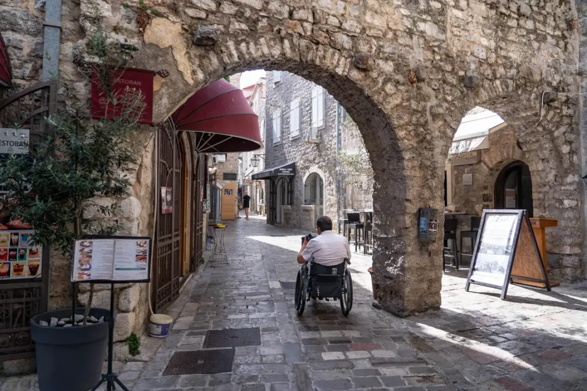 Wąska, brukowana uliczka w **Budva stare miasto**. Mężczyzna na wózku inwalidzkim przejeżdża pod kamiennym łukiem. Po bokach sklepy i restauracje.