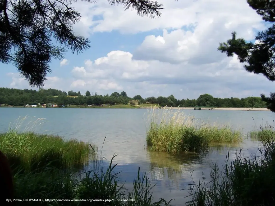 Spokojny zalew Dźibice otoczony zielenią, z kempingiem na brzegu i chmurami na niebie.