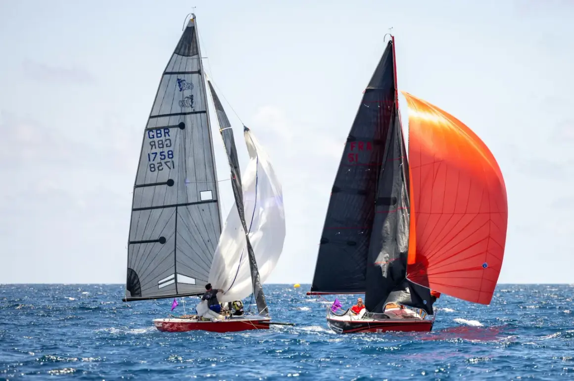 Due barche a vela competono in mare aperto. La barca a sinistra ha una vela grigia con numeri e simboli, mentre quella a destra sfoggia vele nere e arancioni. Lo strallo, elemento cruciale per la stabilità, è ben visibile.