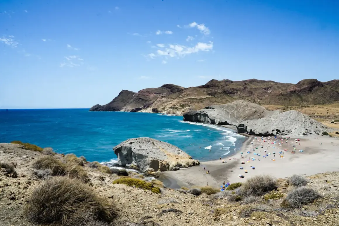 Vista de una playa con gente disfrutando del sol y el mar azul, con formaciones rocosas y montañas al fondo. A lo lejos se divisa el faro de los genoveses.
