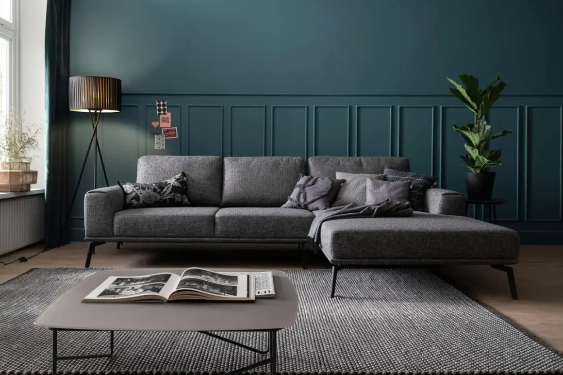 Ein moderner Wohnbereich mit einem grauen Ecksofa, das auf einem gemusterten Teppich steht. Ein Couchtisch mit einem offenen Buch und eine Stehlampe vervollständigen das Bild.