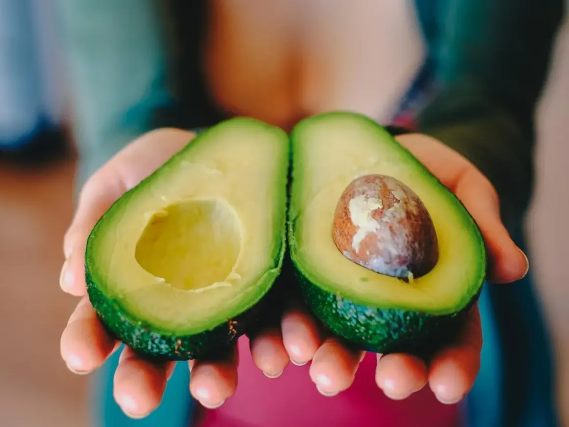 Manos sostienen un aguacate cortado por la mitad, listo para comer aguacate todos los días.