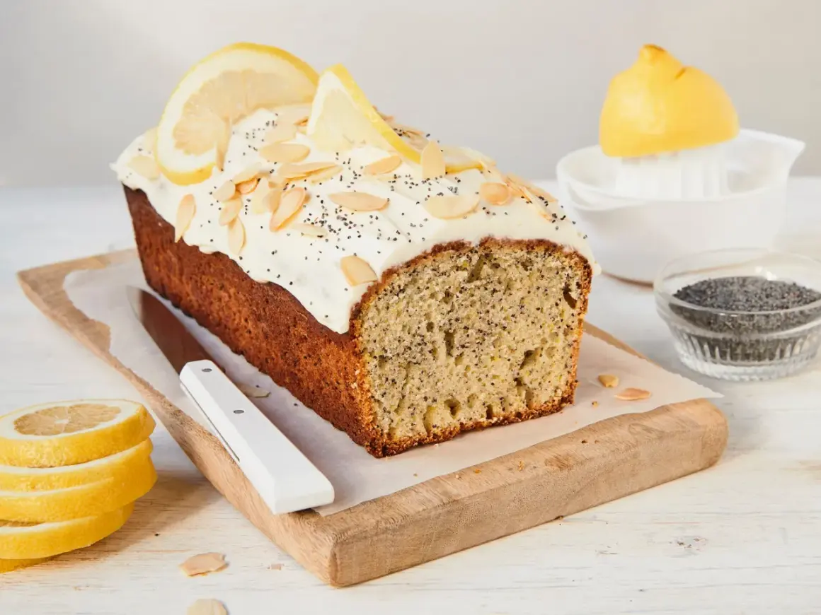 Ein saftiger Zitronen Mohn Kuchen mit weißem Zuckerguss, Mandelsplittern und Zitronenscheiben. Daneben liegen frische Zitronenscheiben und Mohn.