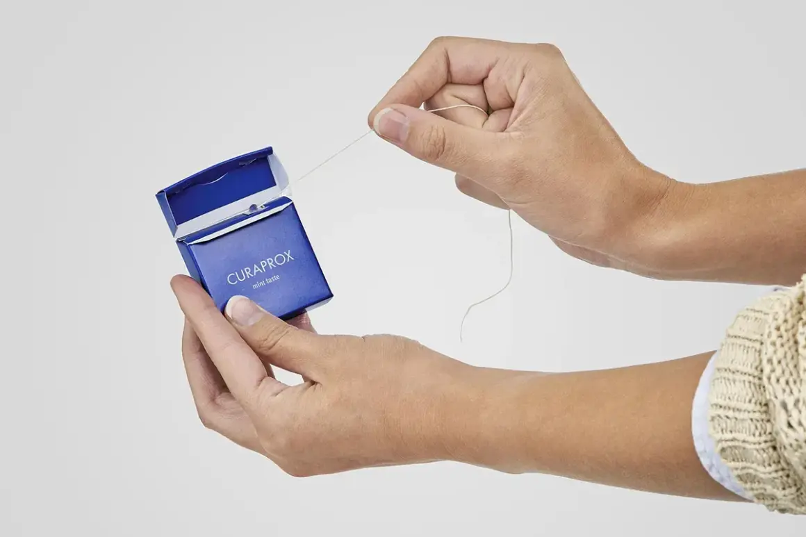 Manos sacando hilo dental de caja azul Curaprox. ¿El hilo dental se pasa antes o después del cepillado?