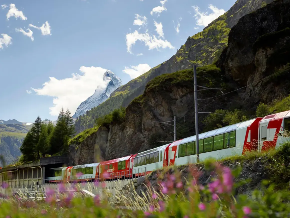 Czerwono-biały pociąg Glacier Express mija skaliste zbocze górskie z ośnieżonym szczytem w tle.