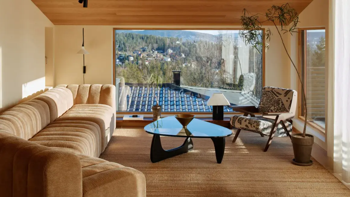 Gemütliches Wohnzimmer im midcentury stil mit einem geschwungenen Sofa, einem Glas-Couchtisch und einem Sessel mit Muster. Große Fenster bieten Blick auf eine bewaldete Hügellandschaft.