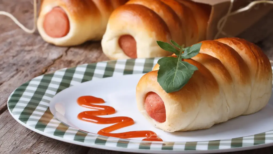 Leckere Bratwurst im Schlafrock, garniert mit einem Blatt, auf einem Teller mit Ketchup-Spur.