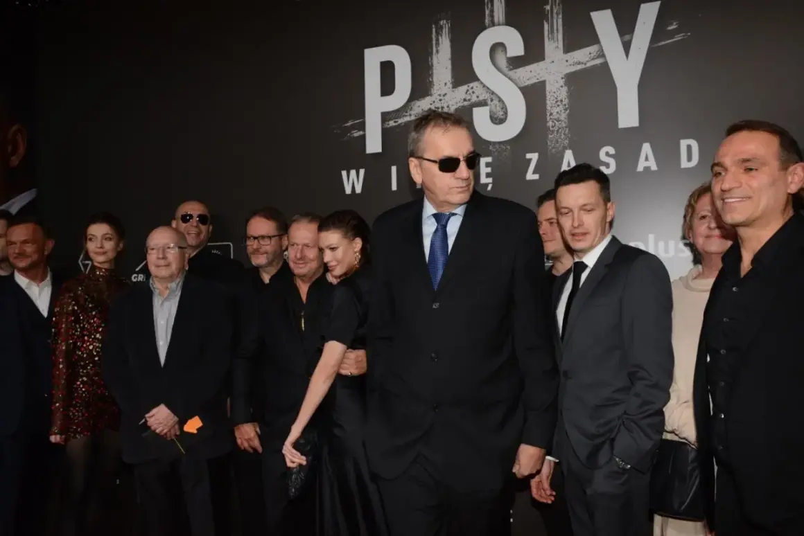 Grupa aktorów z filmu "Psy" pozuje do zdjęcia. W centrum mężczyzna w okularach przeciwsłonecznych i garniturze.