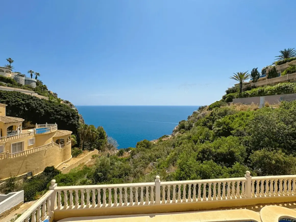 Vista desde una terraza con barandilla blanca hacia el mar azul, con villas en laderas verdes. La puerta del mar San José se abre a un paisaje idílico.