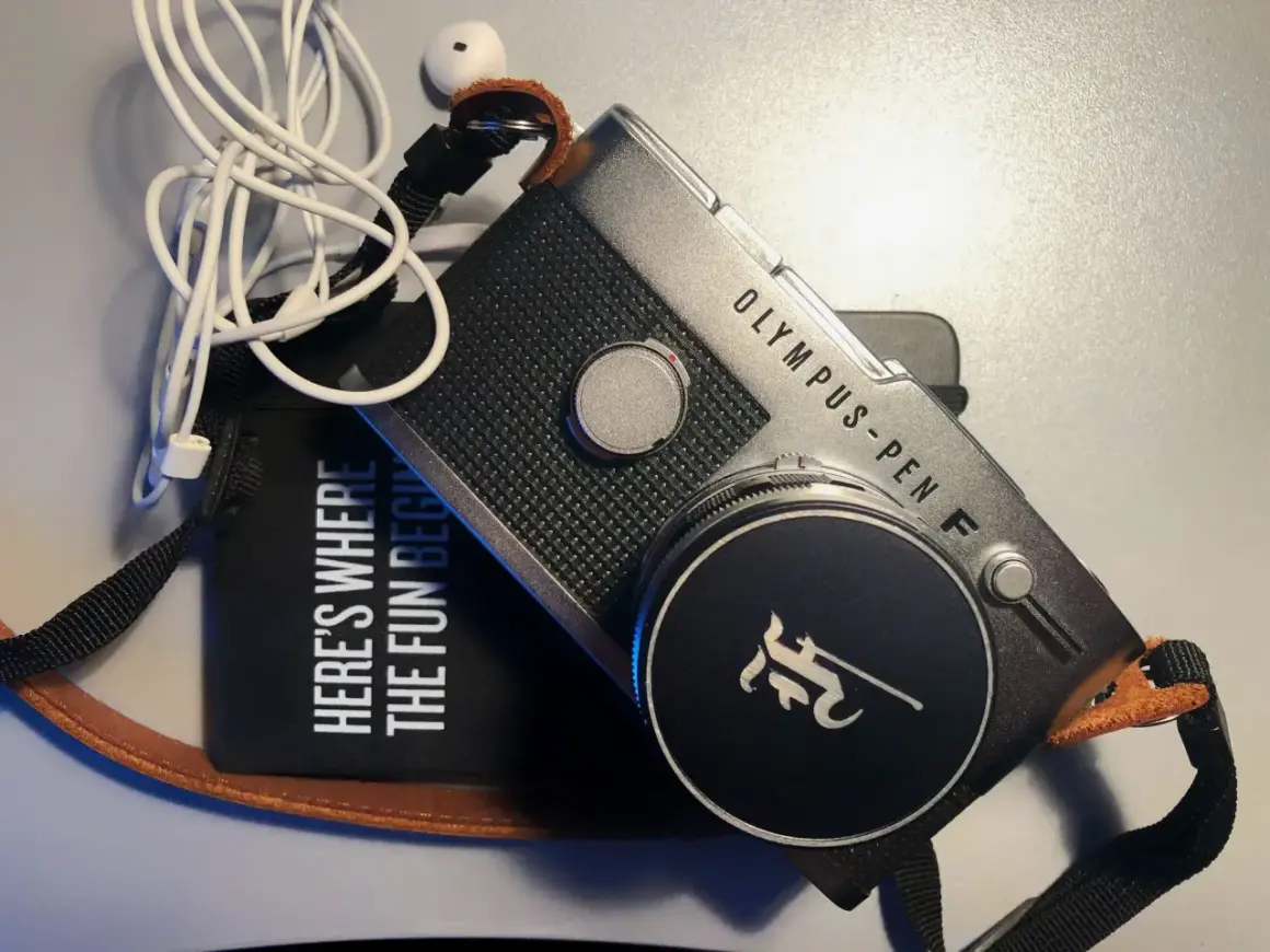 The Olympus Pen FV camera, with its iconic design, sits beside a black pouch that reads "HERE'S WHERE THE FUN BEGINS." White earbuds are tangled nearby.