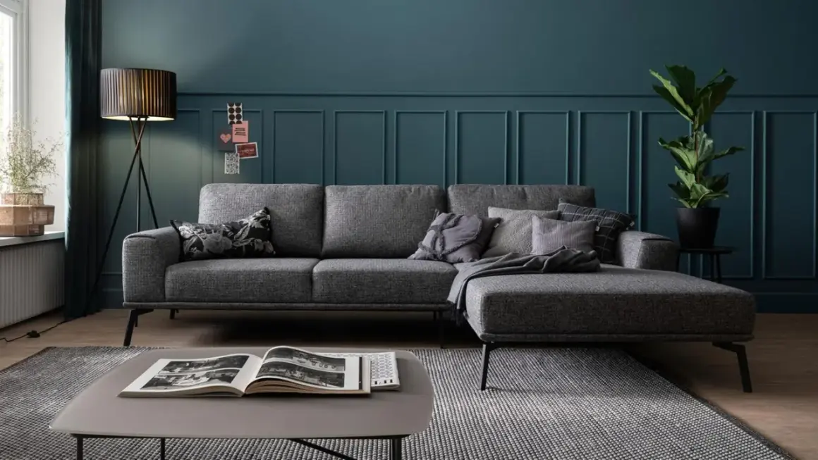 Ein modernes Wohnzimmer mit einem grauen Sofa, das mit Kissen und einer Decke dekoriert ist. Ein Stehlampen-Design und eine Zimmerpflanze runden das Bild ab.