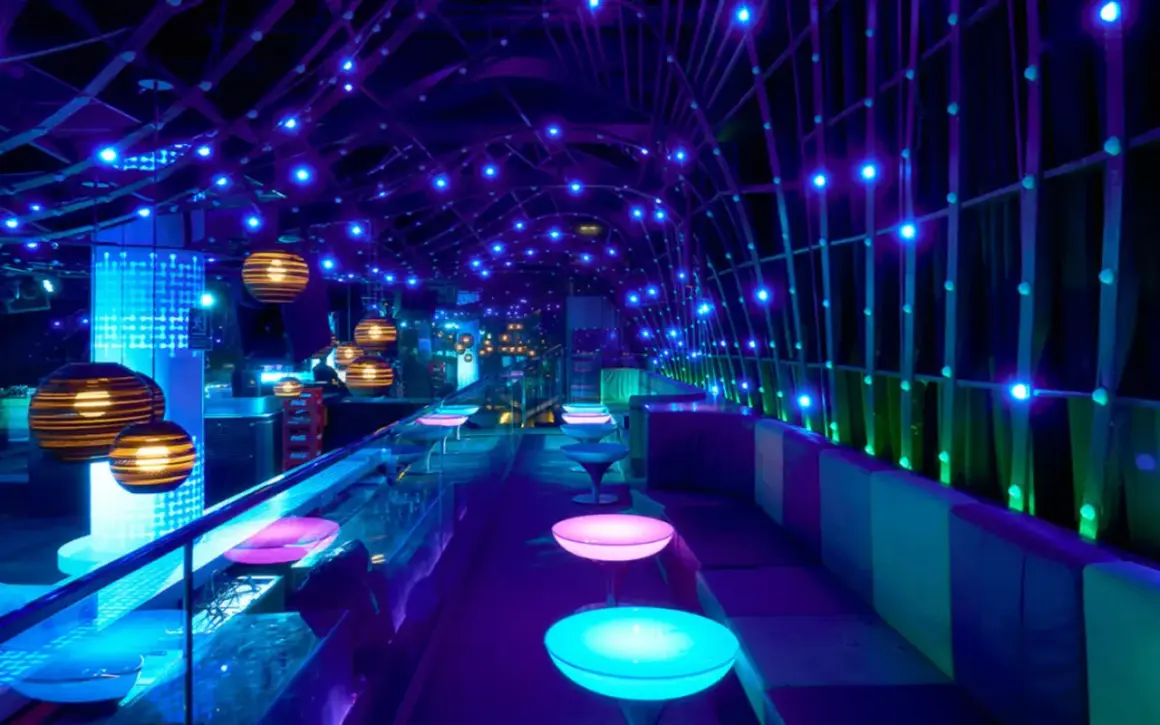 Interni di un night club a Valencia, con luci blu e viola, tavoli luminosi e sedute comode.