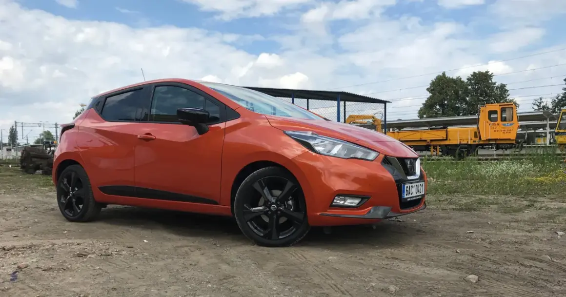 Pomarańczowy Nissan Micra na tle torów kolejowych. Sprawdź nissan micra wymiary, by poznać jego kompaktowe gabaryty.