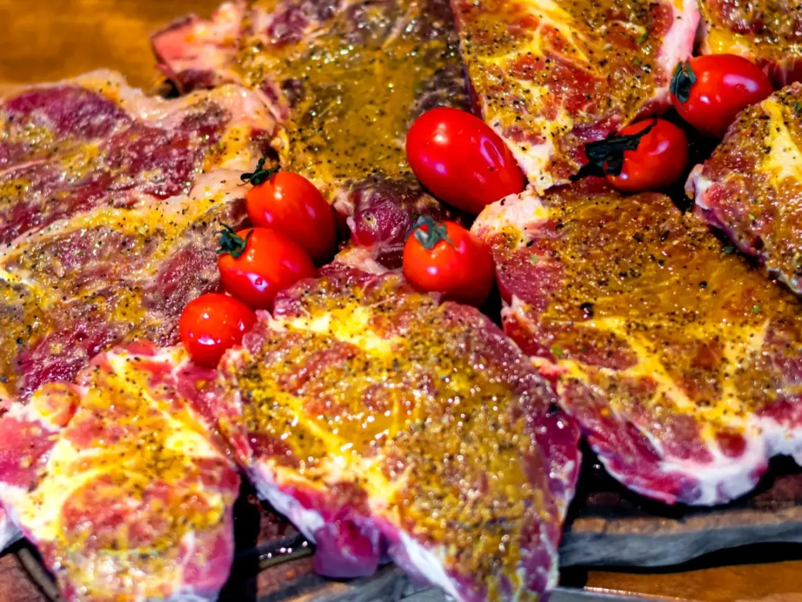 Soczyste plastry karkówki, marynowane i przyprawione, gotowe do grillowania. Obok leżą czerwone pomidorki koktajlowe.
