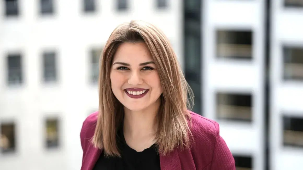 Lächelnde junge Frau mit braunen Haaren, die einen pinken Blazer trägt. Sie studiert IU Master Wirtschaftsinformatik und blickt optimistisch in die Zukunft.