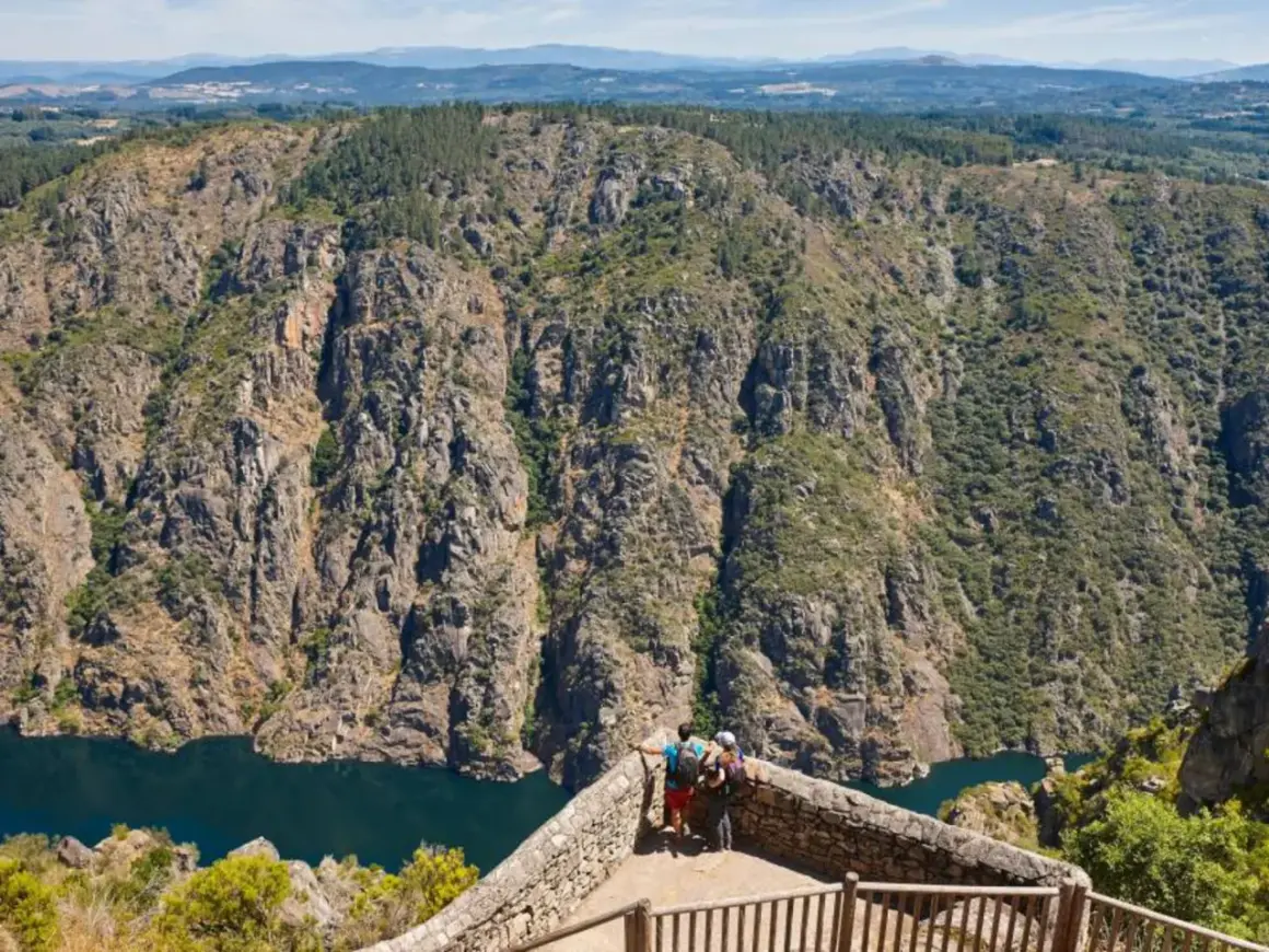 Dos excursionistas contemplan la majestuosidad de los ríos de Galicia, serpenteando entre cañones rocosos y bosques frondosos.