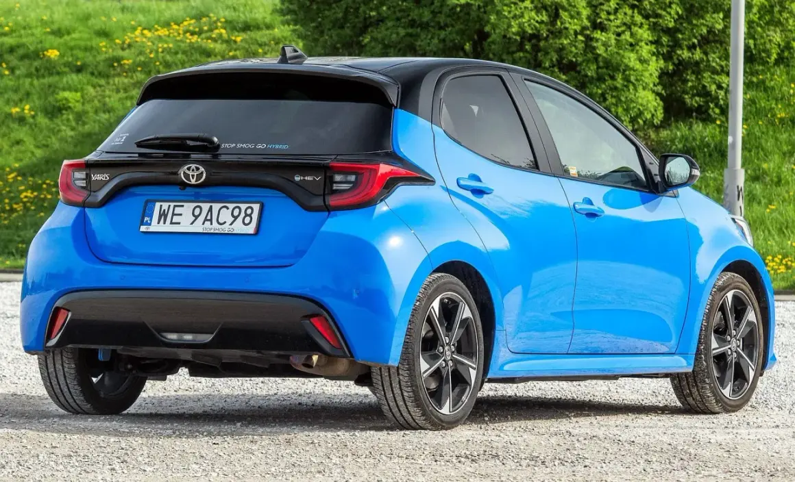 Niebieski Toyota Yaris IV z czarnym dachem, zaparkowany na żwirowej drodze, z zielonym tłem.