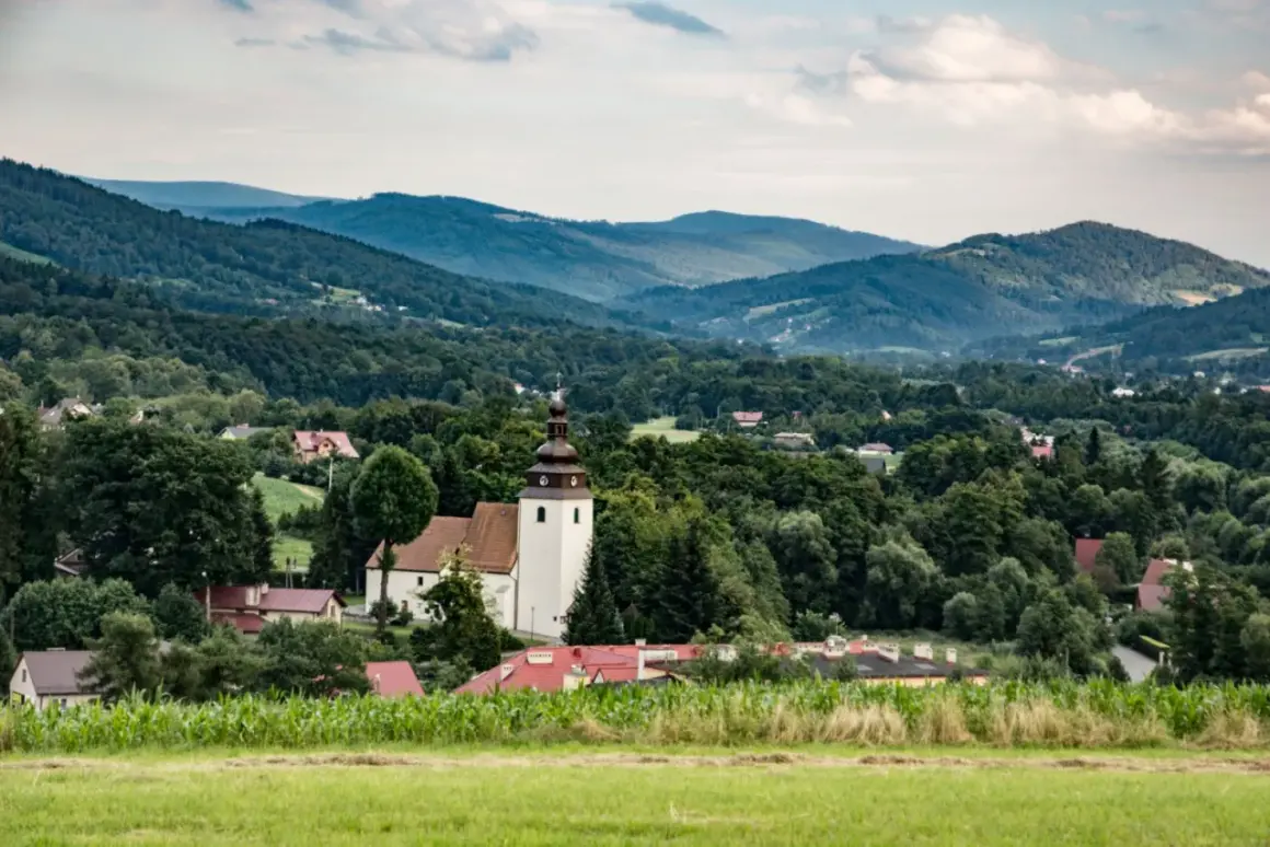 Widok na wieś z kościołem i domami, otoczoną przez zielone wzgórza i lasy. W oddali widać łagodne **górki wielkie**.