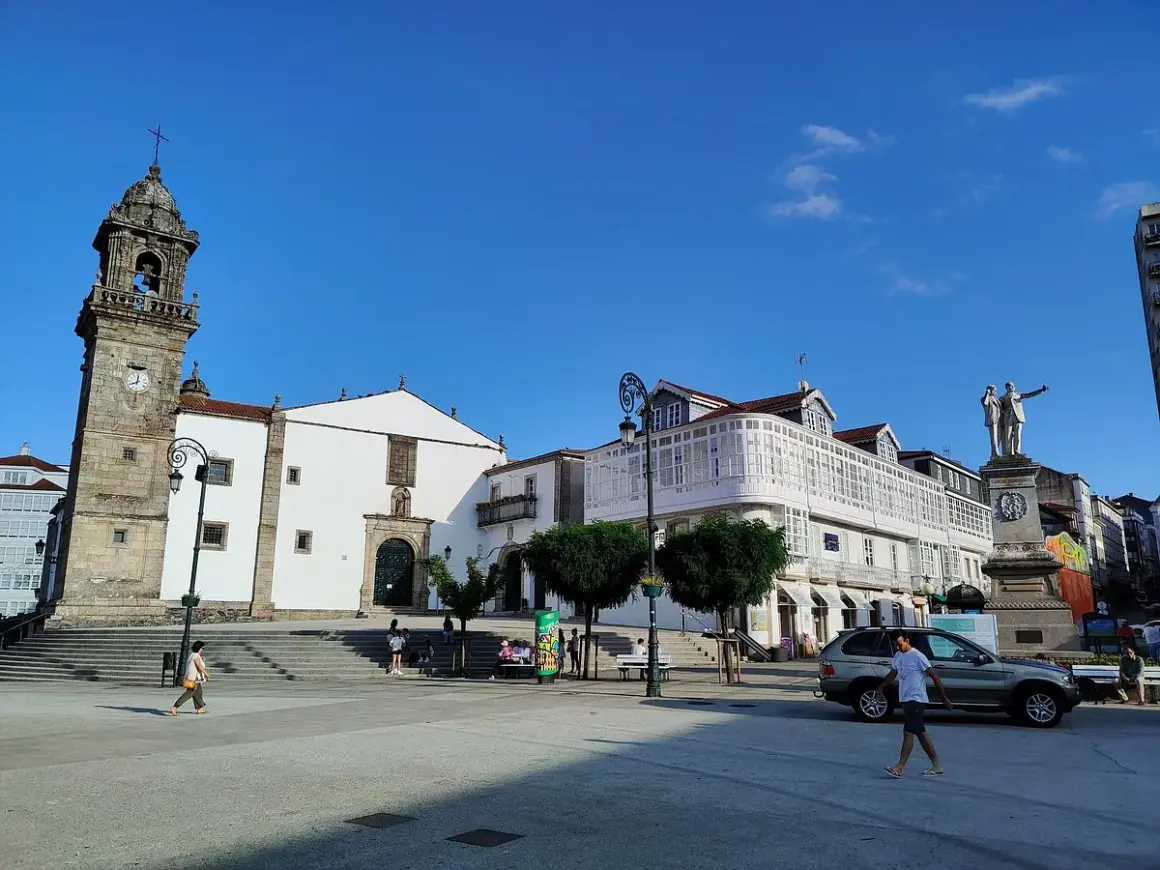La plaza Betanzos, con su iglesia de torre campanario, edificios blancos y un monumento, bajo un cielo azul.
