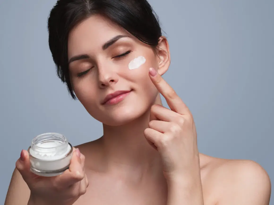 Frau trägt eine Tages- und Nachtcreme auf. Sie hält ein Glas mit der Creme in der Hand und tupft sie mit dem Finger auf ihre Wange.