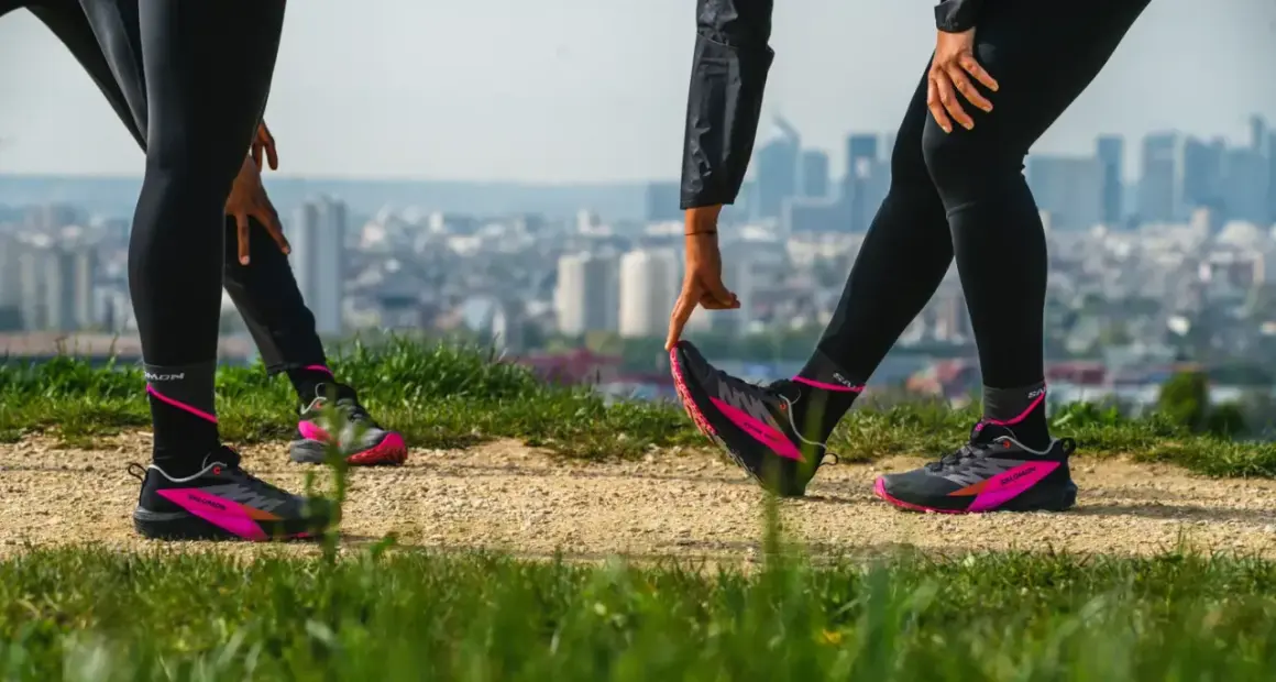Zwei Läuferinnen dehnen sich auf einem Hügel mit Blick auf eine Stadt. Sie tragen schwarze Laufhosen und auffällige pink-schwarze Laufschuhe. Ein Zeichen für den Wiedereinstieg ins joggen nach Geburt.