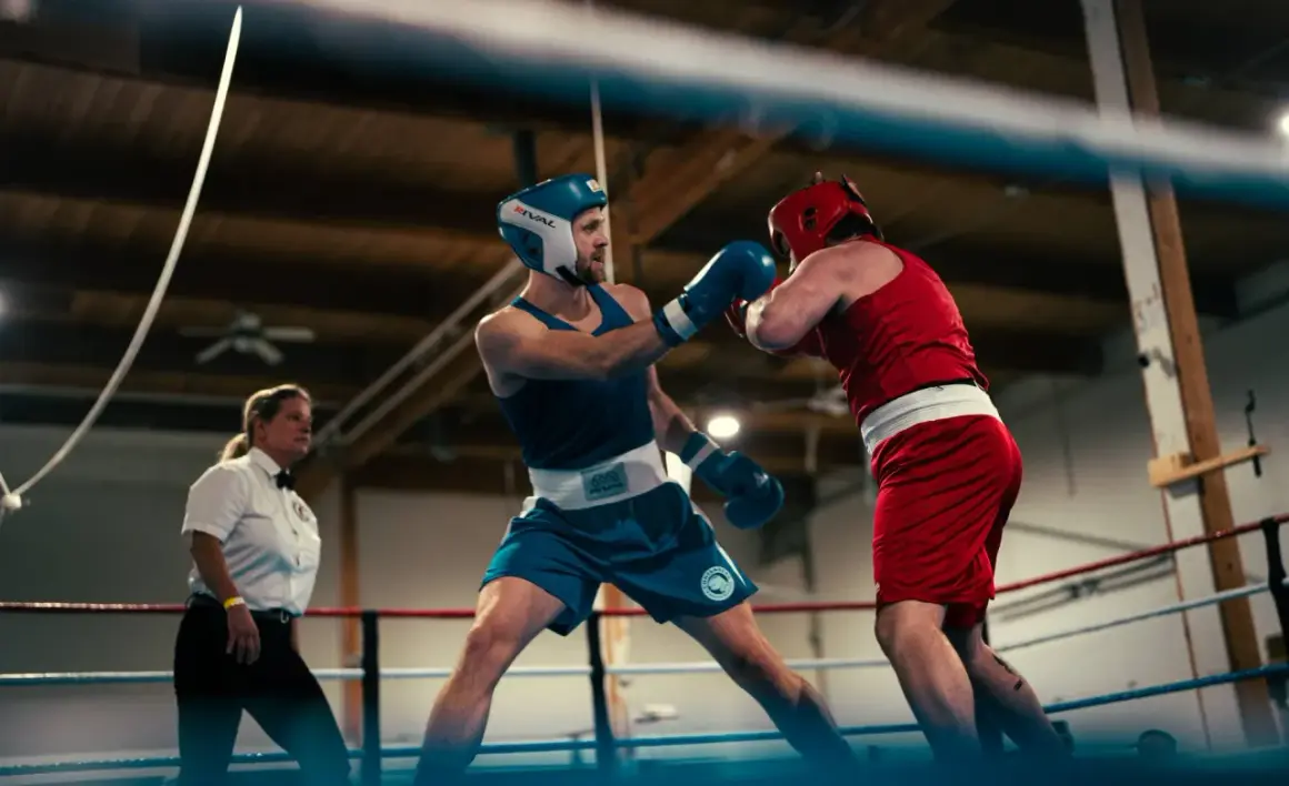 Two boxers in a ring, one in blue with a strong punching stance, the other in red. A referee watches intently.