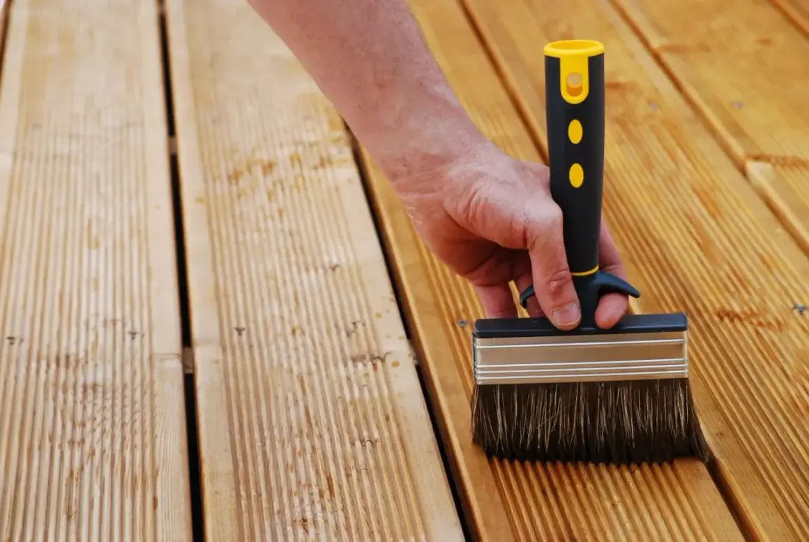 Main d'une personne utilisant une large brosse pour nettoyer bois après ponçage. La terrasse en bois est prête pour le traitement.