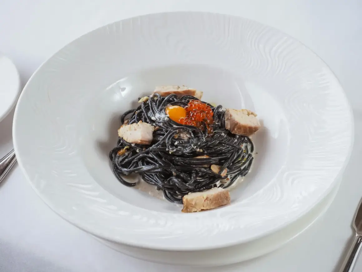 Pasta negra con trozos de pescado, huevas de salmón y yema de huevo. La tinta de calamar, ingrediente principal, aporta un sabor único y propiedades beneficiosas.